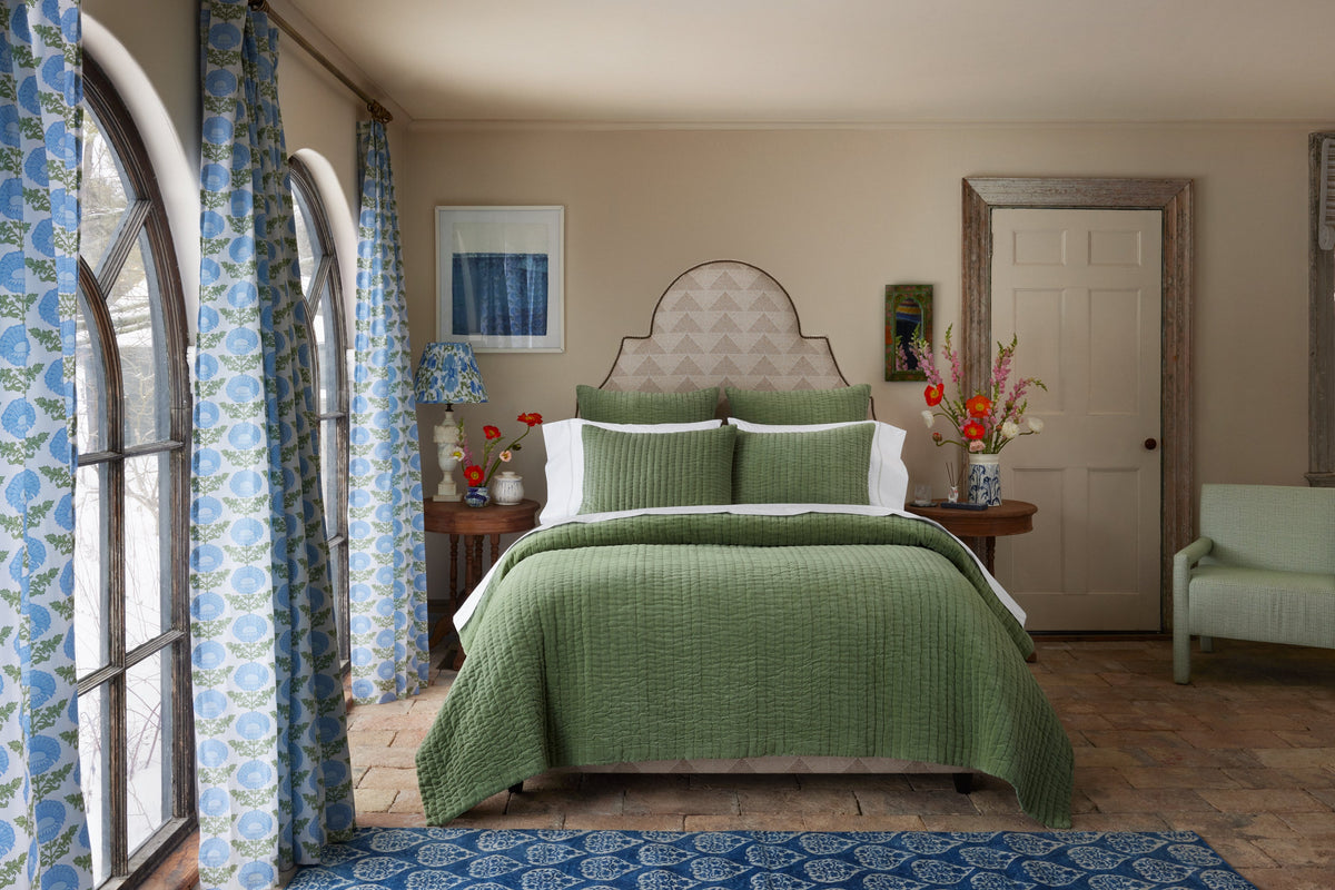A bed with a green quilted coverlet and shams, centered in a room with arched windows and blue floral curtains.