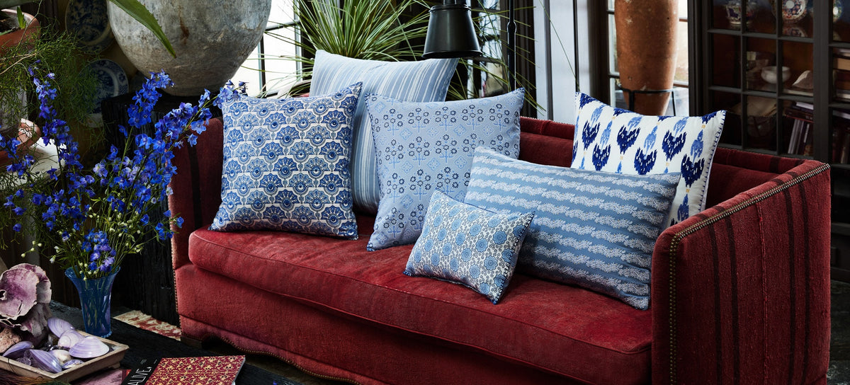 A red velvet sofa with five patterned blue and white throw pillows is set in a room with plants, blue flowers, and decorative items.
