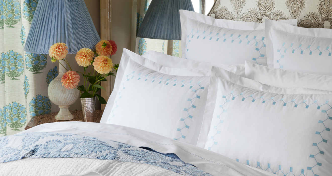 A neatly made bed with white linens featuring light blue geometric embroidery, decorative pillows, a blue patterned throw, and a bedside table with a lamp and vase of orange and yellow flowers.