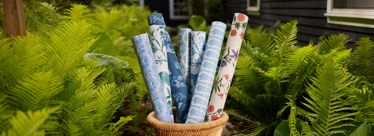 A wicker basket holds several rolls of patterned wallpaper with blue, white, and floral designs, surrounded by green ferns in a garden setting.