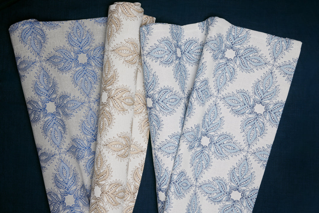 Three folded pieces of fabric with intricate leaf and floral patterns in blue and beige tones, displayed on a dark background.