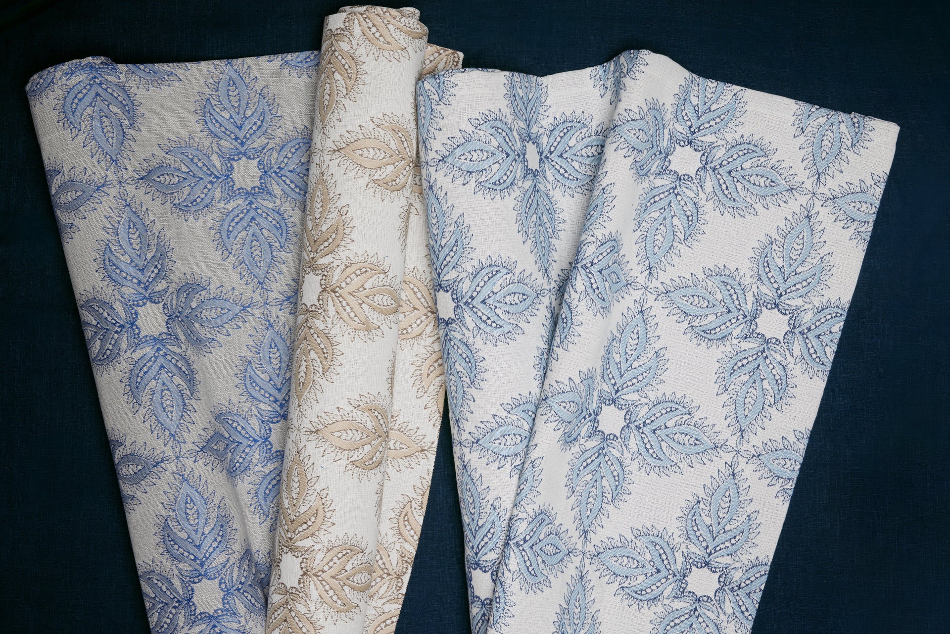 Three folded pieces of fabric with intricate leaf and floral patterns in blue and beige tones, displayed on a dark background.