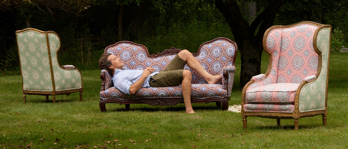 A man in casual clothes lies on an ornate sofa outdoors, with two matching armchairs positioned on grass nearby. Trees and greenery are visible in the background.