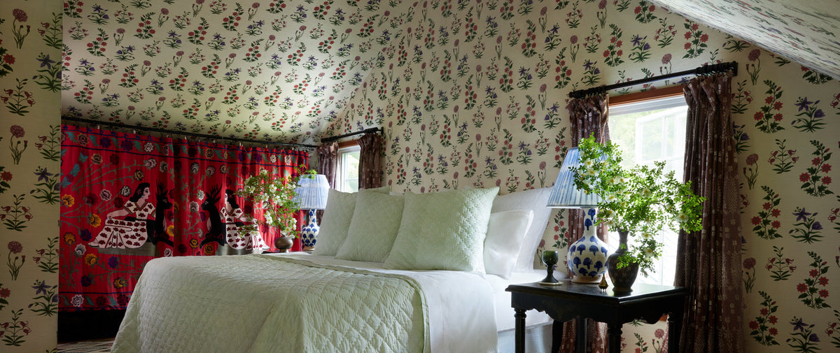 Bedroom with floral wallpaper, a white bed, two side tables with blue and white lamps, and a red tapestry with figures on the left wall. Two windows with brown curtains bring in natural light.