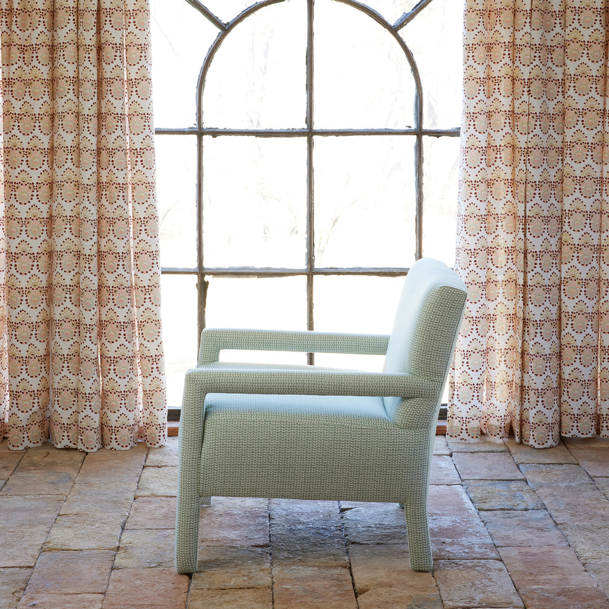 Placed before a large arched window with patterned curtains and set on a tiled floor, the Square Chair in Aleppo Sage by John Robshaw features custom upholstery in durable performance fabric—an elegant seating choice that brings both comfort and style to your living space.