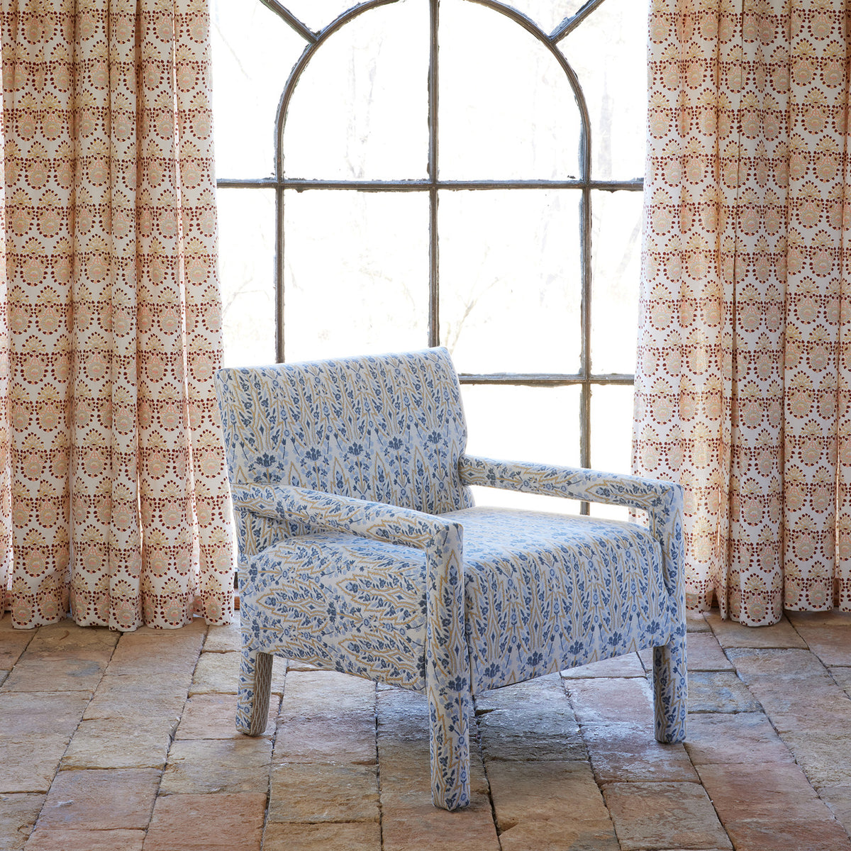 The Square Chair in Marmar Marigold and Natesh Sand by John Robshaw, upholstered in a vibrant performance fabric, sits on a tiled floor in front of a tall arched window with patterned curtains.
