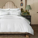 Bedroom with a neatly made bed featuring the John Robshaw Nandi Dark Sage Organic Sheet Set, patterned headboard, bedside table with lamp, floral arrangement, and framed decor on a beige wall.