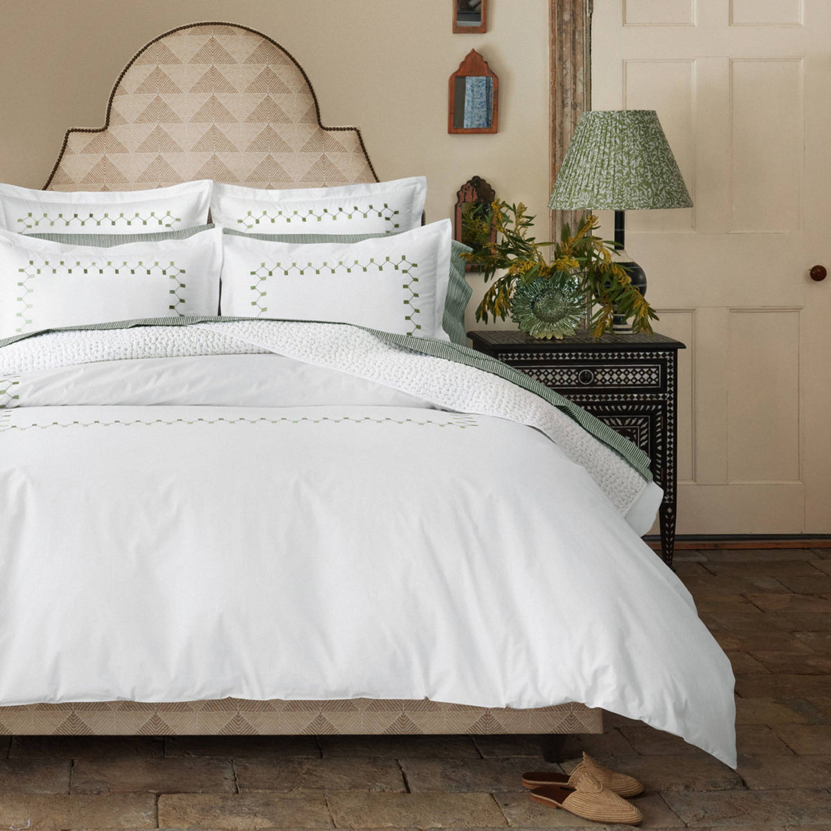 Bedroom with a neatly made bed featuring the John Robshaw Nandi Dark Sage Organic Sheet Set, patterned headboard, bedside table with lamp, floral arrangement, and framed decor on a beige wall.