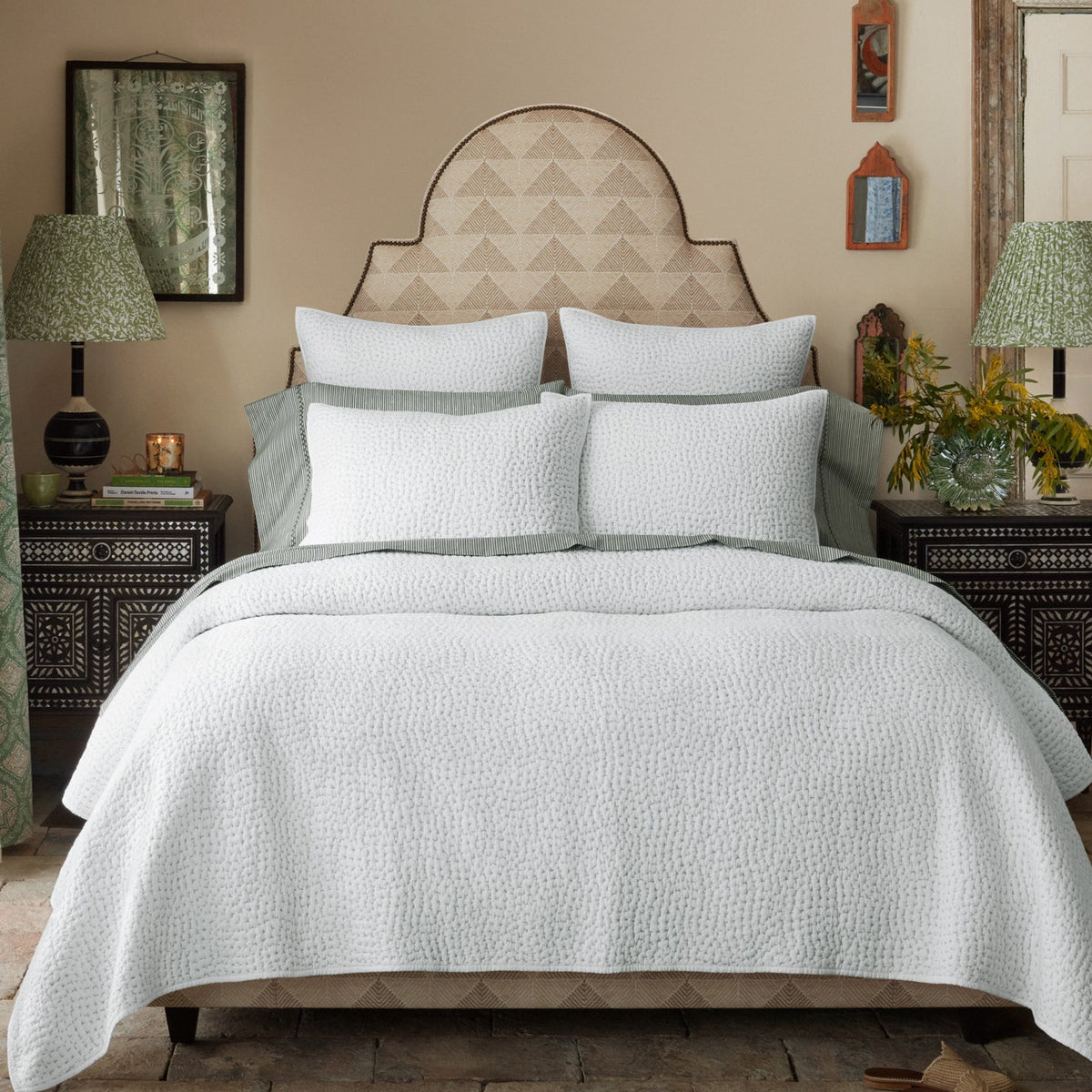 A neatly made bed with white textured bedding, four pillows, and two patterned lamps on dark bedside tables is accented by contemporary decorative items on beige walls; the modern feel is elevated by John Robshaw’s Farzu Sand Performance Fabric, featuring subtle geometric prints.
