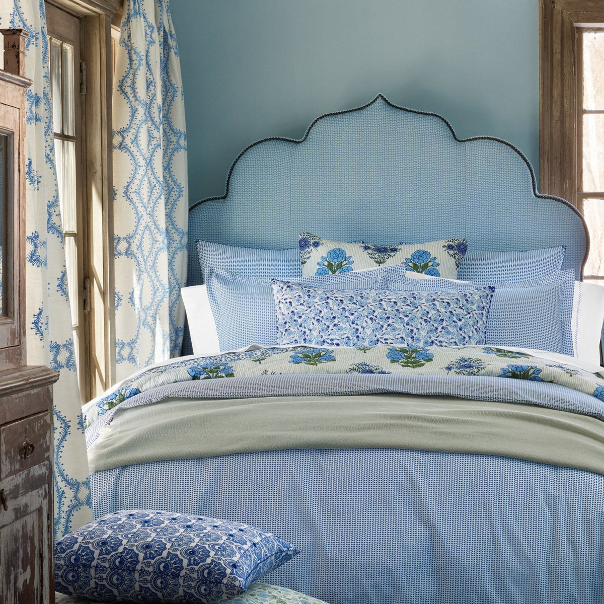 A neatly made bed with a scalloped blue headboard, blue and white floral bedding, and matching curtains in a cozy room with light blue walls and wooden window frames. The Ipsita Indigo Bolster by John Robshaw adds an artisanal touch, perfectly complementing the coastal India vibe of the space.