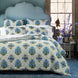 A bed with the Anketi Navy Organic Sheet Set by John Robshaw stands before a blue headboard, near a window and a table holding a vase of purple flowers.
