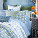 A neatly made bed features the John Robshaw Cinde Light Indigo Organic Sheet Set, complemented by blue and green patterned pillows and bedding, and sits beside a gray nightstand adorned with a vase of orange flowers.