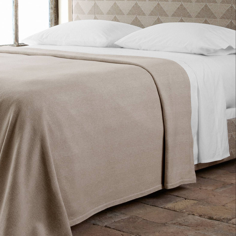 A bed with the Anketi White Organic Sheet Set by John Robshaw, white pillows, and a beige blanket sits on rustic tiles with a patterned headboard in the background.