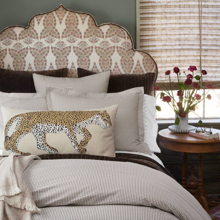 A neatly made bed with patterned bedding, a cheetah pillow, and the John Robshaw Velvet Coffee Quilt. A round table holds red flowers and a book by a window with bamboo shades.