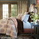 A sunlit room with French doors features a neatly made bed with floral bedding, brown and white pillows, and the John Robshaw Velvet Coffee Quilt beside a blue nightstand with flowers.