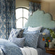 A bed featuring a patterned light blue headboard, blue and white pillows and bedding crafted from elegant Divit Willow Fabric by John Robshaw, complemented by floral curtains and a round window behind, with a vase of flowers resting on a nearby table.