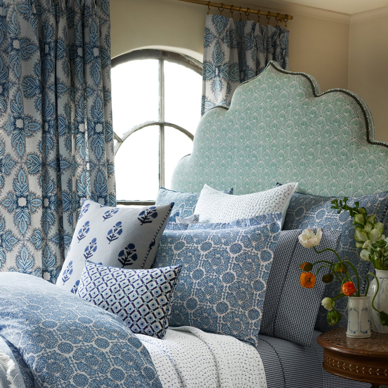 A bed featuring a patterned light blue headboard, blue and white pillows and bedding crafted from elegant Divit Willow Fabric by John Robshaw, complemented by floral curtains and a round window behind, with a vase of flowers resting on a nearby table.