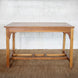 The Taru Teak Table by John Robshaw, hand crafted with a rectangular solid teak top and simple, straight legs, is displayed on a woven rug against a white brick wall.