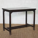 The Chhaya Teak Table by John Robshaw, a handcrafted rectangular dark wood piece featuring four straight legs and a sturdy stretcher base, sits atop a woven mat against a white brick wall, radiating the timeless charm of vintage furniture.