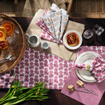 A table set for breakfast includes pastries, fruit, cutlery, a cup, saucer, glass, and John Robshaw's Sabah Berry Napkins (Set of 4) featuring block printed floral patterns on handmade cotton placemats under natural light. - 31263989563438