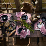 A rustic dining table set with black plates, Nandi Berry Napkins (Set of 4) by John Robshaw made from 100% cotton, various foods including bread and cheese, candles, and greenery. Sunlight filters in through large windows in the background. - 31263958302766