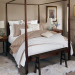 A neatly made bed with white and beige Nivi Euro bedding by John Robshaw, a wooden four-poster frame, a bench at the foot, side tables with lamps, artwork on the wall, an embroidered pillow featuring natural linen fabric, and a small vase with flowers. - 31263122194478