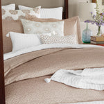 A neatly made bed with the John Robshaw Nivi Euro beige patterned comforter and decorative pillows, including an embroidered pillow, is set in a bedroom with ambient decor like books, a lamp, and a vase of flowers. The bedding features hidden zipper closures for a seamless look. - 31263122161710
