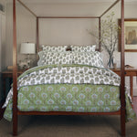 A neatly made four-poster bed with floral-patterned bedding in green and white, showcasing the Nilay Leaf Organic Duvet by John Robshaw, a nightstand with a lamp, and decorative branches in a vase. - 31249535172654