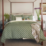 A well-made bed with the Atulya Moss Duvet Set from John Robshaw, featuring green patterned bedding, a decorative throw, and a pillow with a colorful design sits between two nightstands. The cotton linen duvet adds to the cozy setup, with each nightstand holding plants and various items. - 31249530781742