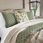 A neatly made bed with a green patterned comforter and matching pillows, accented by the John Robshaw Farah Marigold Decorative Pillow featuring a lemon motif. A vase of purple flowers and artwork are in the background. - 31259421671470