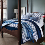A neatly made bed with a dark wooden frame, adorned with white and blue patterned bedding and multiple pillows, including the embroidered Zain Kidney Pillow from John Robshaw. A lamp and framed art can be seen on a side table by the window in the background. - 31263930187822