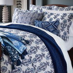 A neatly made bed with a white and blue floral bedspread, matching pillows, and a dark blue textured blanket. There are additional decorative pillows with geometric and floral designs, including the embroidered Zain Kidney Pillow from John Robshaw. A lamp is on the side table. - 31263930220590
