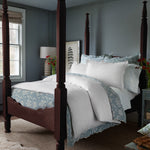 A sophisticated bedroom featuring a wooden four-poster bed, adorned with the John Robshaw Amina Ecru Duvet Set, crafted from the finest quality cotton linen. A nightstand with a lamp, flowers in a vase, and a window with light streaming in complement the light blue walls. - 31249526915118