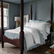 A bedroom featuring a dark wood four-poster bed adorned with the Kimari Mist Coverlet by John Robshaw in light blue and white jacquard cotton chenille, accompanied by a nightstand with a lamp, and vases of delicate woven florals on each side. The walls are blue-gray with a framed artwork near the bed.