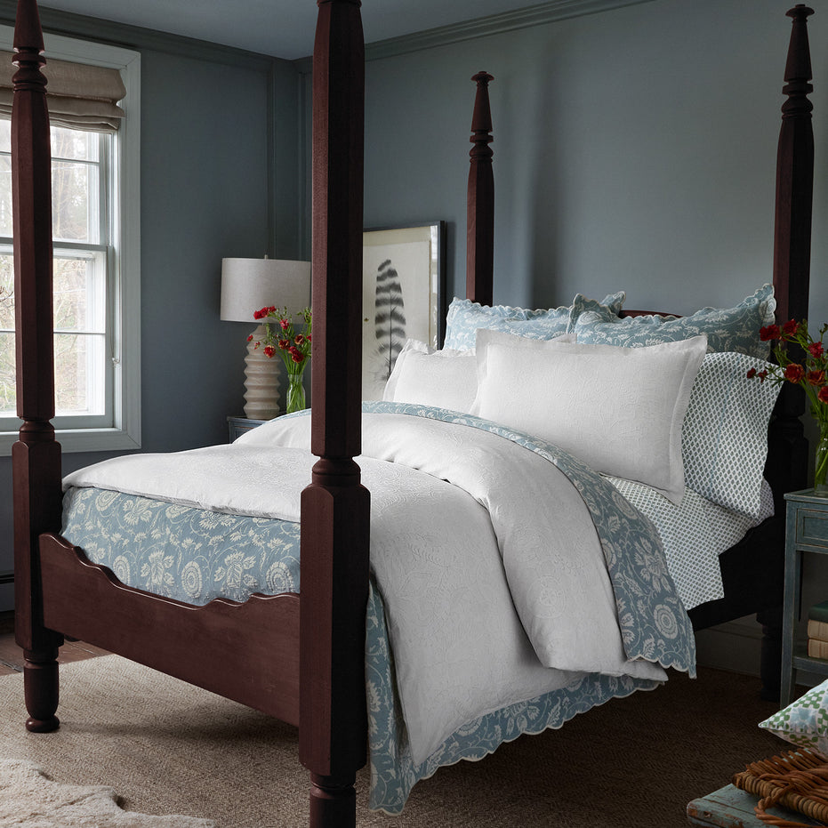 A bedroom featuring a dark wood four-poster bed adorned with the Kimari Mist Coverlet by John Robshaw in light blue and white jacquard cotton chenille, accompanied by a nightstand with a lamp, and vases of delicate woven florals on each side. The walls are blue-gray with a framed artwork near the bed.