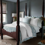 A bedroom featuring a dark wooden four-poster bed adorned with the luxurious Amina Ecru Duvet Set by John Robshaw, showcasing the finest quality cotton linen in white and blue hues. Nearby, a bedside table displays flowers alongside a lamp, while a window with blinds offers light control. The walls are painted in a serene muted blue-grey tone.
 - 31249526882350