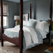 A neatly made four-poster bed with the John Robshaw Kimari Mist Coverlet, featuring delicate woven florals in blue and white, is centered in a room with light gray walls. A window with a beige shade and a small nightstand adorned with a lamp and flowers complete the serene setting.
