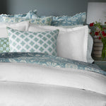 A neatly made bed adorned with multiple pillows in various blue and white patterns, covered by the finest quality cotton linen from the Amina Ecru Duvet Set by John Robshaw, and a lamp with red flowers on a nearby nightstand. - 31249526849582