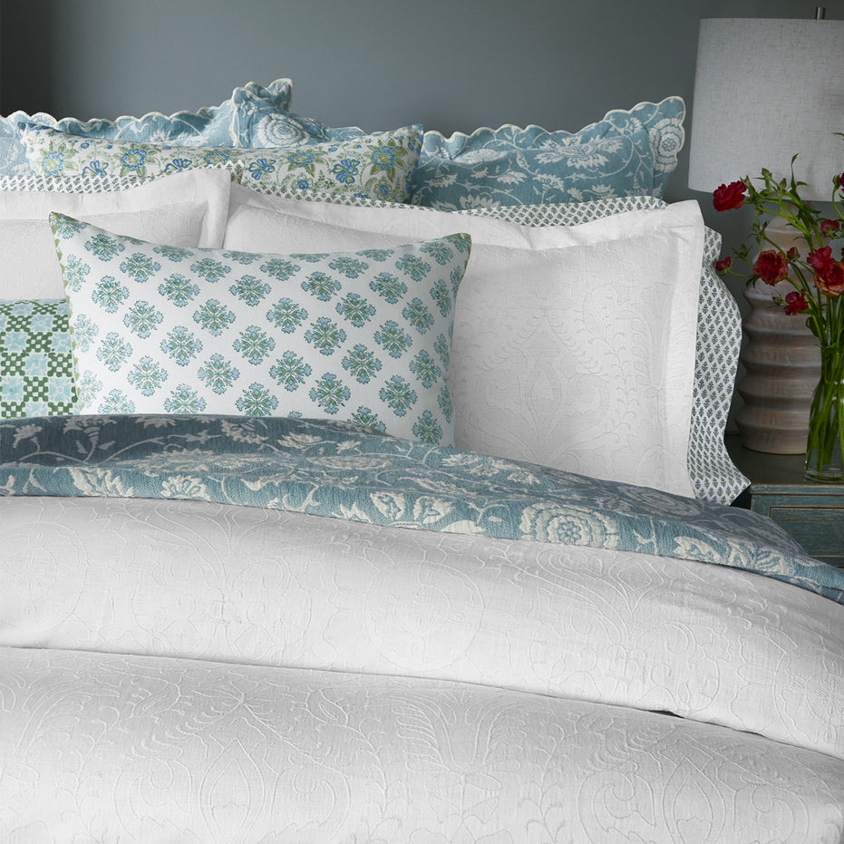 A neatly made bed adorned with the Kimari Mist Coverlet by John Robshaw, crafted in soft jacquard cotton chenille with intricate scalloped edges, complemented by multiple pillows in various shades of blue and green. Nearby stands a bedside table holding a stylish lamp and a vase filled with vibrant red flowers.