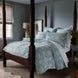 A bedroom featuring a four-poster bed adorned with the Kimari Mist Coverlet from John Robshaw, showcasing blue floral bedding made of soft jacquard cotton chenille. Bedside tables flank the bed, each holding a vase of flowers. In the background, a window decorated with a roman shade and framed artwork contribute to the room's delicate charm.