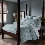 A bedroom featuring a four-poster bed adorned with the Kimari Mist Coverlet from John Robshaw, showcasing blue floral bedding made of soft jacquard cotton chenille. Bedside tables flank the bed, each holding a vase of flowers. In the background, a window decorated with a roman shade and framed artwork contribute to the room's delicate charm. - 31258913767470