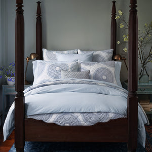 A wooden four-poster bed with light blue stylized print bedding and matching pillows graces a well-lit room. The John Robshaw Cinde Light Indigo Organic Duvet adds a touch of luxury, flanked by nightstands with lamps on each side, and complemented by floral arrangements in the background.