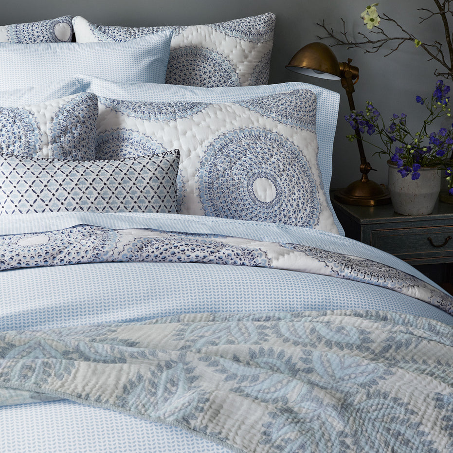 A neatly made bed displays patterned pillows, a quilt with blue and white designs, and the Cinde Light Indigo Organic Duvet by John Robshaw. Beside the bed sits a wooden nightstand adorned with a brass lamp and a vase of flowers.