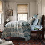 A neatly made bed adorned with the John Robshaw Asma Peacock Woven Quilt, featuring a reversible blue and white floral print, stands near a window with beige blinds. A wooden nightstand holds a glass pitcher, a book, and a potted plant. Branches in a vase are in the corner. - 31258828734510