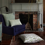 A cozy living room features an Ojas Lavender Euro from John Robshaw, a hand block printed navy blue armchair with patterned cushions, a large floor pillow, a lamp, a brick fireplace with a tool set, and greenery hanging above the mantle. - 31263323619374