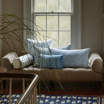 A beige sofa is placed in front of a window adorned with multiple Yavi Azure Bolsters from John Robshaw, each featuring hand-stitched edging. A branch with green leaves extends into the frame. A blue patterned rug with mirror embellishments is positioned on the wooden floor. - 31263913639982