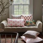 A cozy nook with a small sofa in front of a window, adorned with six Lucy Coral Bolster pillows by John Robshaw, each featuring vibrant floral patterns and colors, all hand block printed and made in India. - 31261340794926