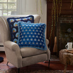 A beige armchair with one cotton velvet indigo pillow and an Isaya Decorative Pillow by John Robshaw sits beside a wooden fireplace with a broom. A wood stool next to the chair holds a white mug and saucer. - 31259460501550