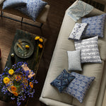Living room scene with a beige sofa adorned with various blue and white patterned throw pillows, including a beautiful embroidered John Robshaw Zain Kidney Pillow. A rustic wooden coffee table holds drinks and yellow flowers, while a nearby stool features additional pillows with Shibori printing. - 31263929597998
