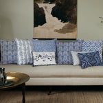 A beige sofa is adorned with a variety of patterned blue and white cushions, including the Esha Indigo Decorative Pillow by John Robshaw, featuring block printing and hand-stitched edging. A brass tray rests on the coffee table in the foreground, and a framed painting hangs on the wall above the sofa. - 31259345518638